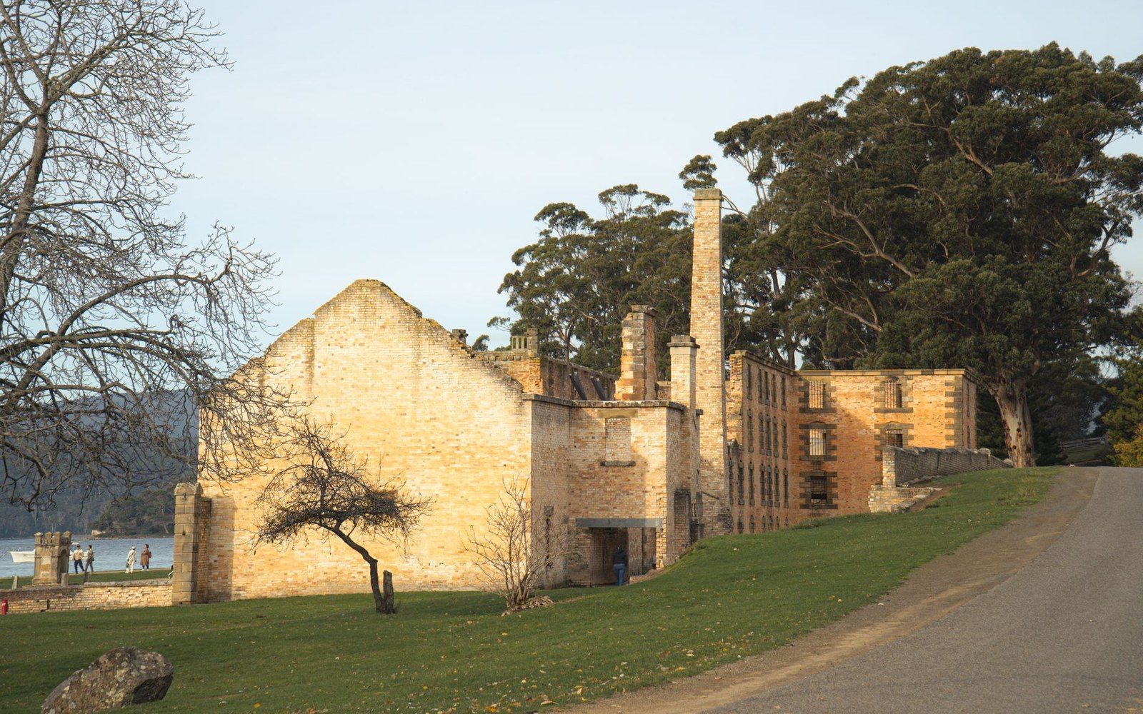 Ruined brick building with tall chimneys stands beside a grassy waterfront lawn